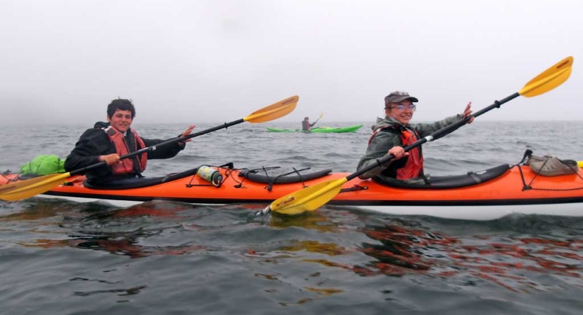 Two people paddle and orange kayak under gray skies. 